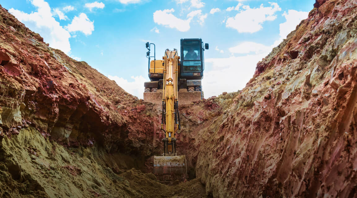 An excavator digging into a large trench cut into reddish rocky earth