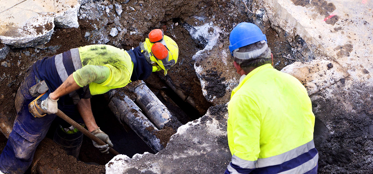 Two field professionals in hardhats and yellow vests examine a large hole dug in a sidewalk to expose pipes beneath