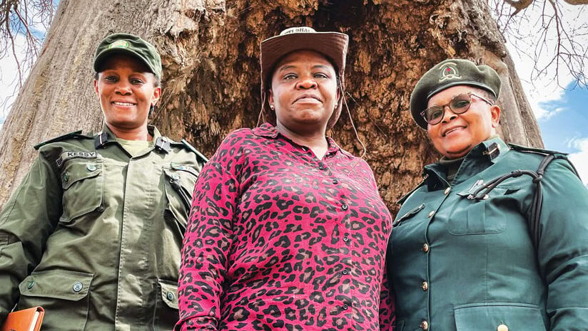 Three women—two in uniforms and one in a pink leopard-print shirt and a hat—standing in front of a large tree, smiling