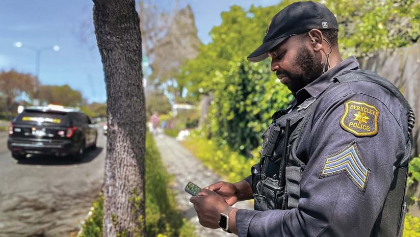 A police officer with a Berkeley Police patch on his uniformed arm standing in a residential neighborhood near a police car looking at a smartphone