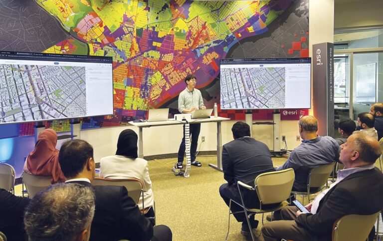 A person standing behind a desk with a laptop on it, flanked by two screens with maps on them, standing in front of a big colorful map, presenting to a room full of people