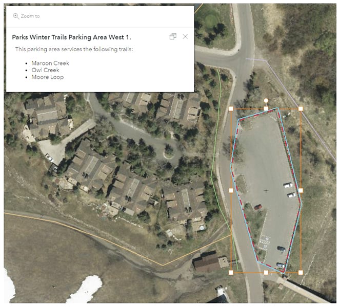 An aerial view of a parking area near some houses with trails marked in tan and green lines and the parking area outlined in orange, blue, and dotted-red polygons