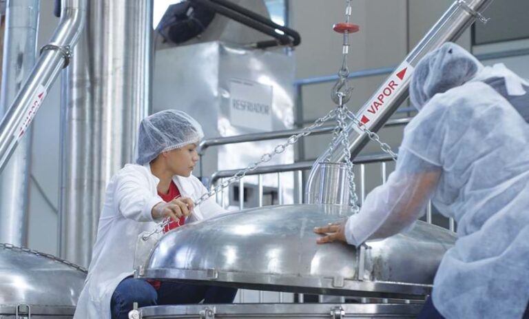 Two people in hairnets and long-sleeve jackets holding down a large lid in a factory