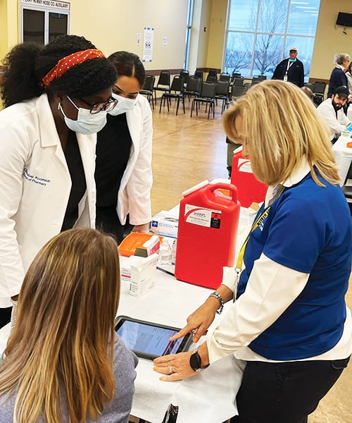 Four women gathered around a table in a large room—two of whom are in lab coats and masks learning how to use the form on a tablet from one of the other women