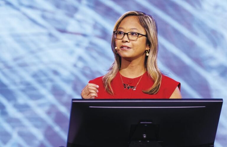 A woman in a red shirt talks onstage from behind a computer monitor