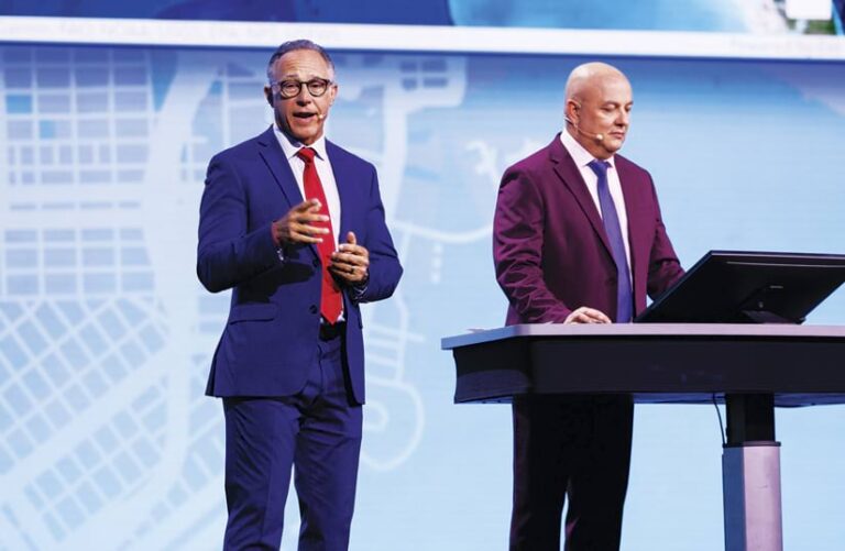 Two men in suits onstage—one talking and one working on a computer