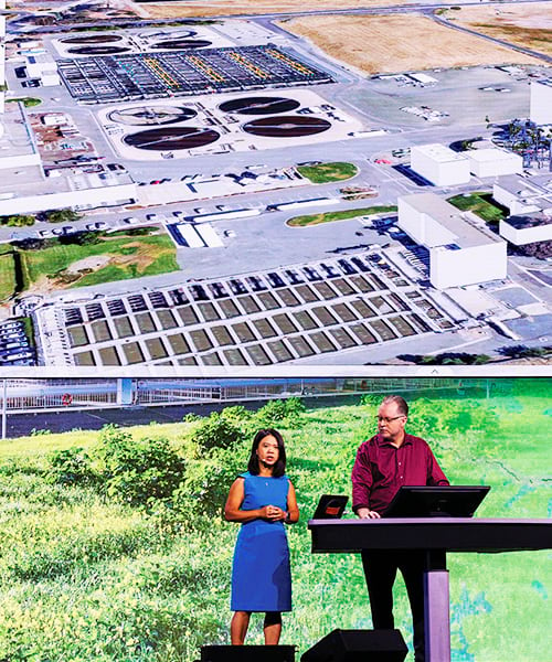 A woman and a man onstage, standing in front of an aerial image of a building, with the woman standing to the left of a table talking while the man works on a computer behind the table