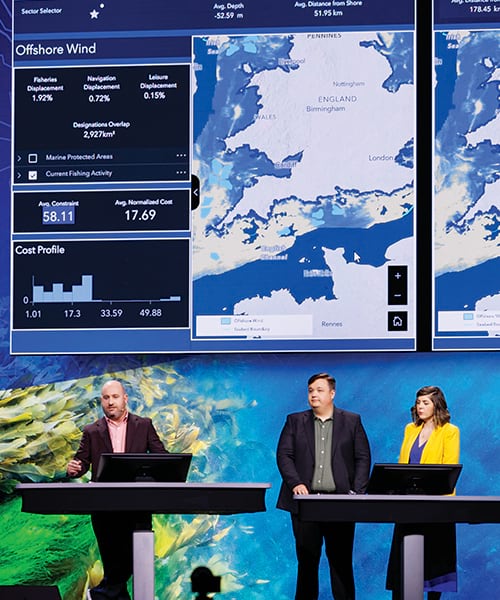 Two men and a woman stand behind computers onstage, presenting in front of a screen that shows maps of England and its surrounding waters