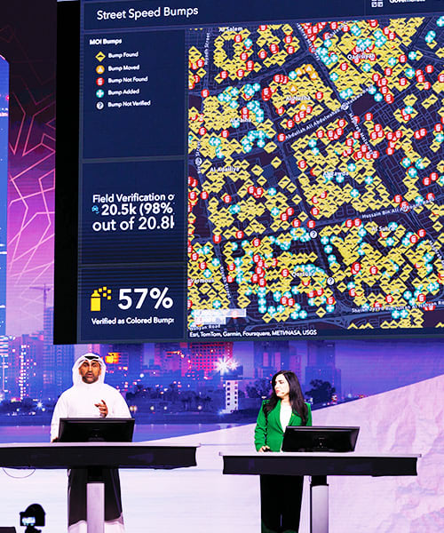 A man and a woman stand onstage behind tables with computers on them, in front of a large screen that shows a dashboard with a map that has yellow, red, and blue location points on it