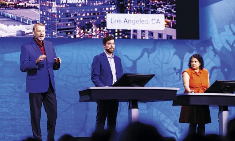 Two men and a woman stand onstage, with one man standing to the left of two tables, talking, and the other two presenters standing behind each table working on computers