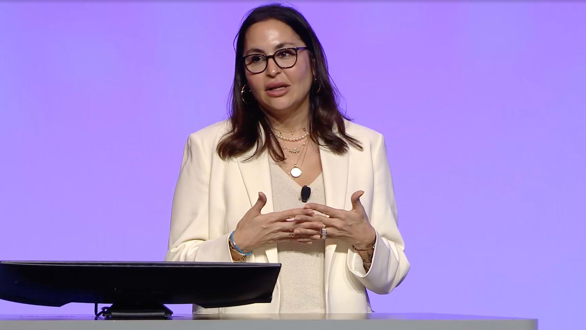 A person wearing a white blazer and blouse standing near a computer in front of a lavender background