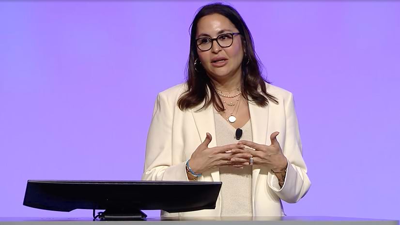 A person wearing a white blazer and blouse standing near a computer in front of a lavender background