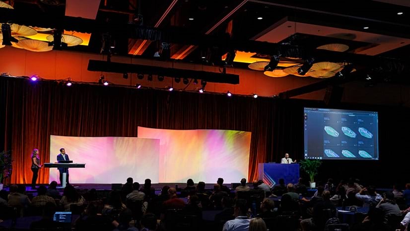 In a darkened room in front of large screens, three presenters address an audience