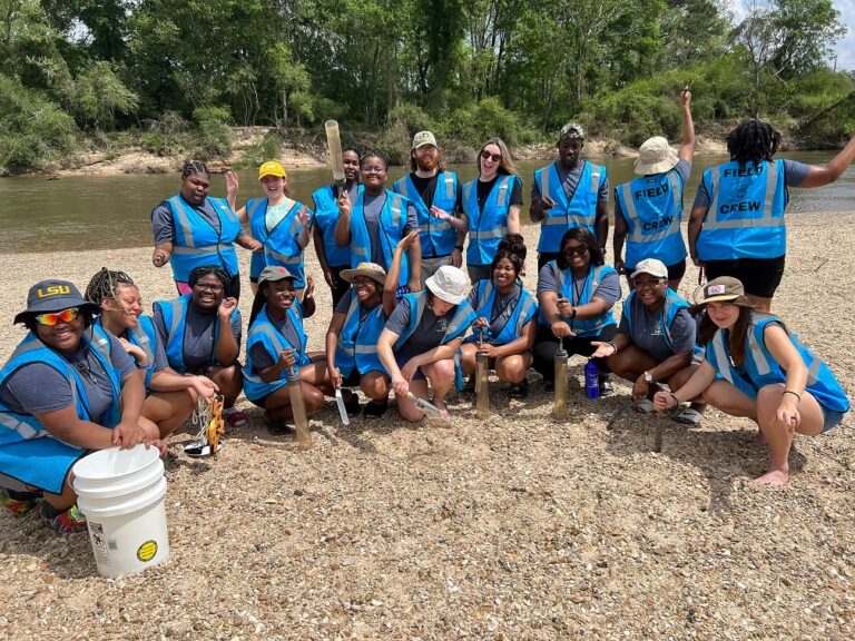 About 20 smiling individuals in blue vests pose in a riparian area