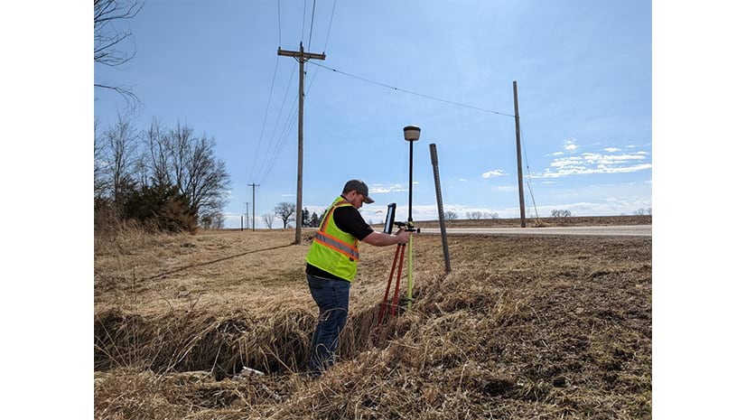 A surveyor in a safety vest stands in a grassy ditch using a GPS receiver mounted on a tripod to take measurements. Power lines and a rural road are in the background.