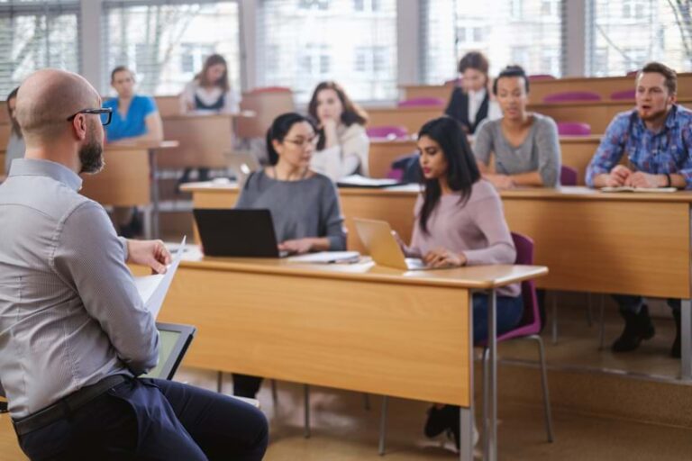 A professor stands in a tiered lecture hall addressing students working at individual desks. The students, who represent diverse ethnicities, are focused on laptops and notebooks.