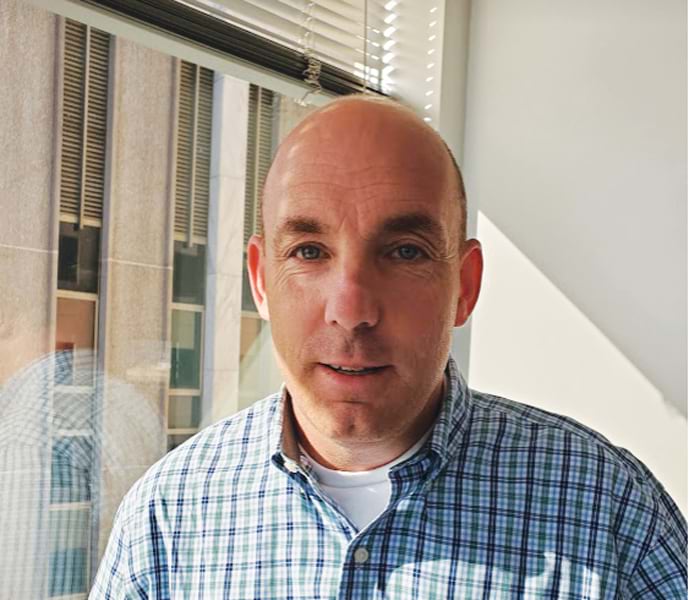 Headshot of a man with short hair and a blue and green plaid shirt, smiling in front of a window with blinds.