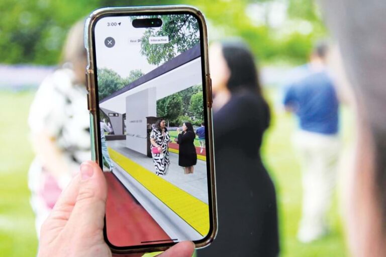 A hand holds a smartphone displaying an augmented reality experience. Two women stand inside a virtual bus station superimposed over a real-world outdoor setting.