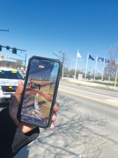 A hand holds up a smartphone displaying an augmented reality-generated transit station in its real-world context. The screen shows a 3D image of a bus station overlaid on the real world as seen through the camera. The background shows a street, trees, and a row of flags.