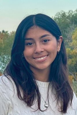 Headshot of a young woman with long, dark hair wearing a white cable-knit sweater. She smiles warmly at the camera. Trees and a blue sky provide a blurred background.