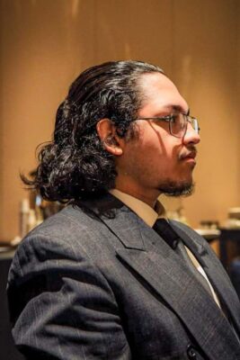 A young adult with long dark hair, glasses, and a goatee wears a dark pinstripe suit jacket with a white collared shirt and dark tie. He is looking to the right of the image.