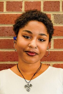 Headshot of a young woman with short curly hair, wearing a white shirt and a dragon pendant necklace. She is standing against a red brick wall and has a neutral expression.