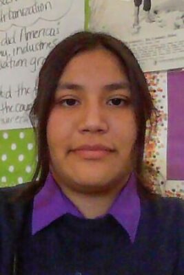 A young woman with brown hair and brown eyes wears a purple collared shirt underneath a navy blue sweater. She stands against a colorful bulletin board covered in text.