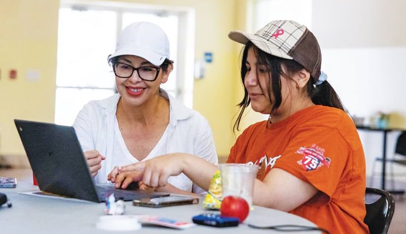 Two women sit at a round table, looking at a laptop in front of them. The woman on the left smiles as she points to something on the screen. The other woman, who wears an orange shirt and plaid baseball cap, looks at the screen with her.