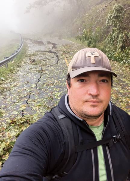 Selfie of a man wearing a baseball cap and backpack on a road through a forest. The road is covered with debris and large cracks from a landslide. The forest is shrouded in fog.