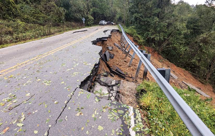 A two-lane asphalt road is crumbling away at the edge where the shoulder should be. The asphalt has fallen away into a ditch with exposed dirt, revealing the road's substructure.