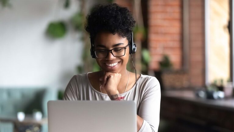 A young woman wearing headphones and glasses smiles at her laptop, her hand resting under her chin.