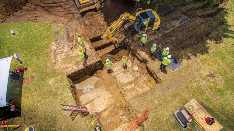 Aerial view of an archaeological dig site. A backhoe rests beside a rectangular pit, with wooden planks laid across sections. Archaeologists in hard hats and safety vests work around the pit.