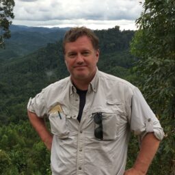 Scott Paul poses in a white button-up shirt in front of a jungle background.