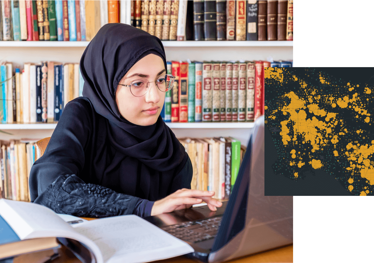 Personne portant un hijab de couleur bleu indigo et des lunettes, utilisant un grand ordinateur posé sur la table d’une bibliothèque scolaire, une pile de livres en arrière-plan
