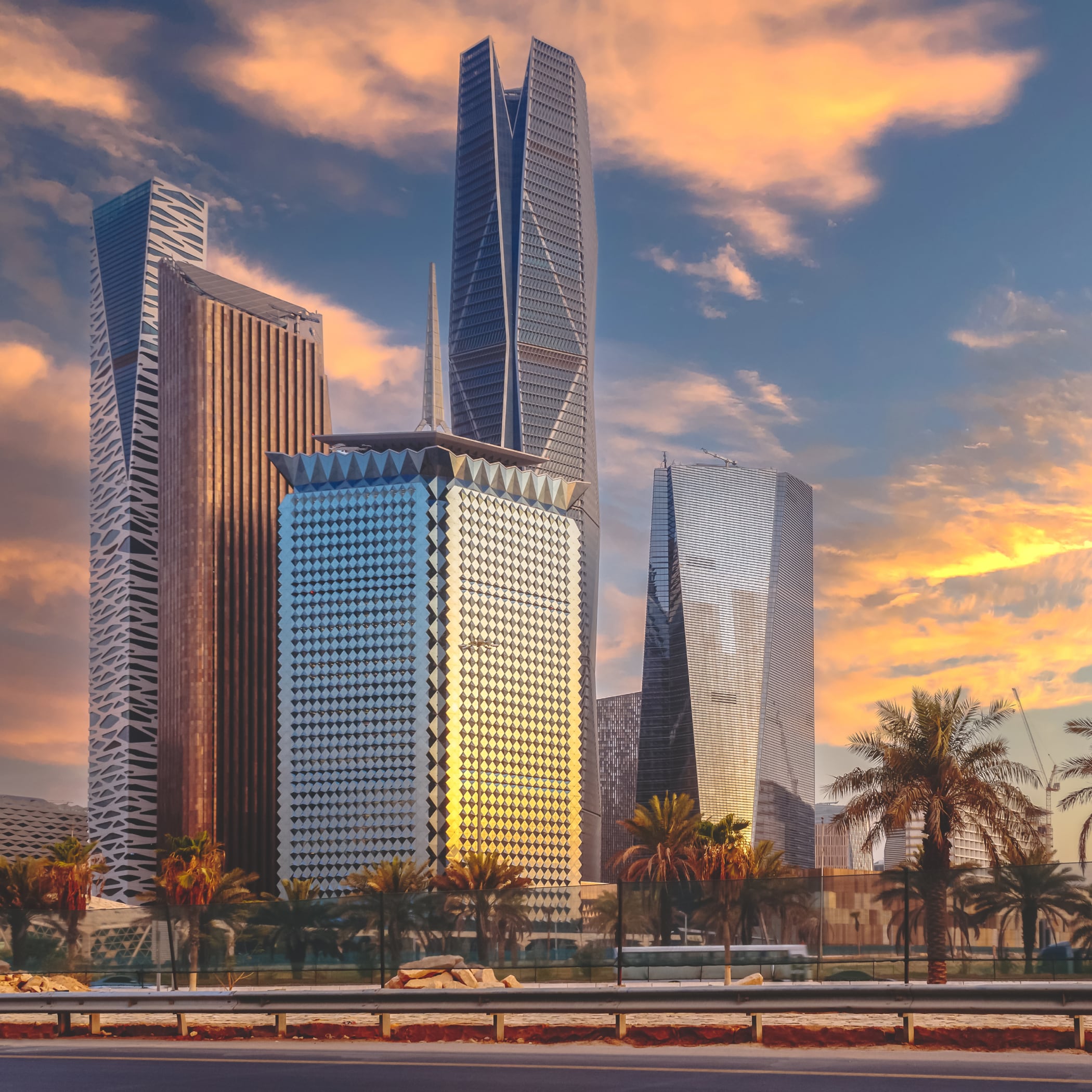 A modern cityscape with soaring skyscrapers surrounded by palm trees under a pale blue sunset sky