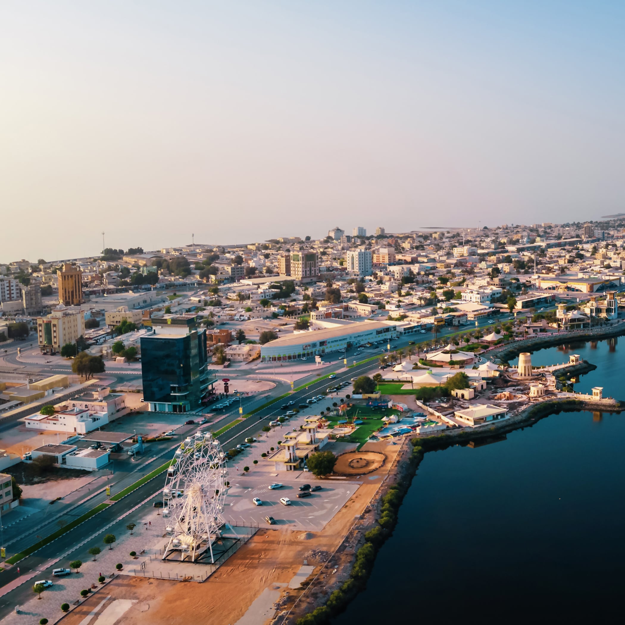 A sprawling modern cityscape beside a still deep blue bay, bordered on one side by a wide swath of green parkland