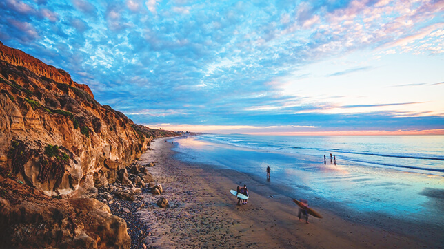 Carlsbad coast line with vibrant blue water, bright orange-red cliffs, and a purple pink sky with clouds.