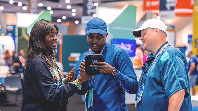 Three conference attendees standing in the exhibit hall looking at a tablet being held by the middle person