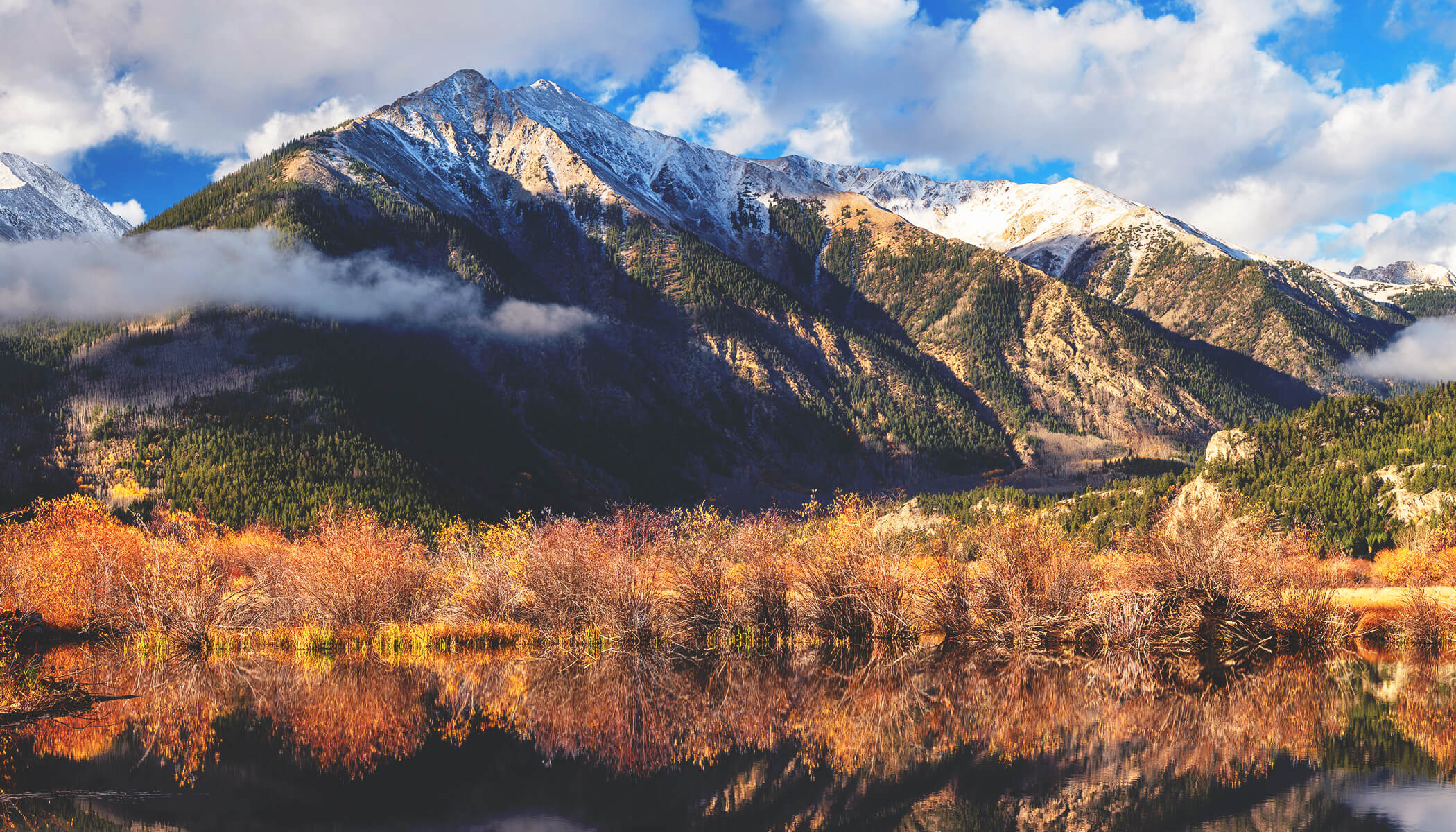 A lake surrounded by brush at the foot of a majestic, snow-capped mountain range 