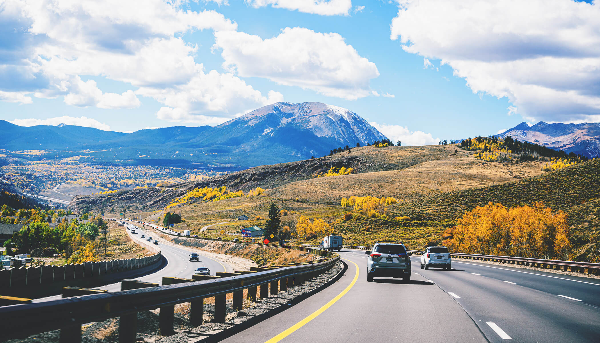 A multilane highway surrounded by rolling, fall-colored hills with a mountain in the distance on a sunny day