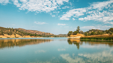 A body of water surrounded by rolling foothills under and blue sky 