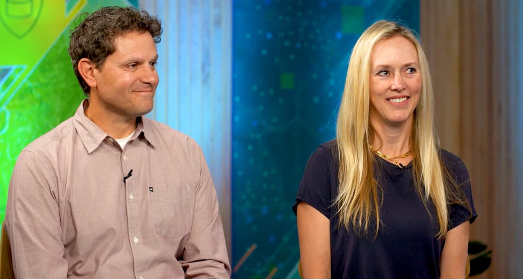 Two Esri employees, one in a light brown button-down shirt and the other in a dark blue T-shirt, are seated side by side against a background of teal, green, and blue graphics