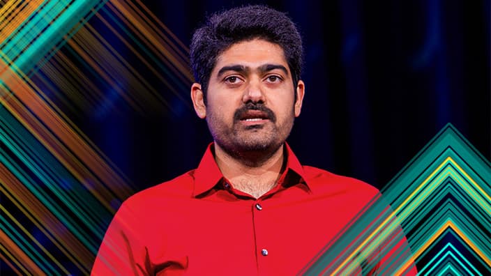 Head and shoulders portrait of a presenter with dark hair and facial hair wearing a vibrant red dress shirt