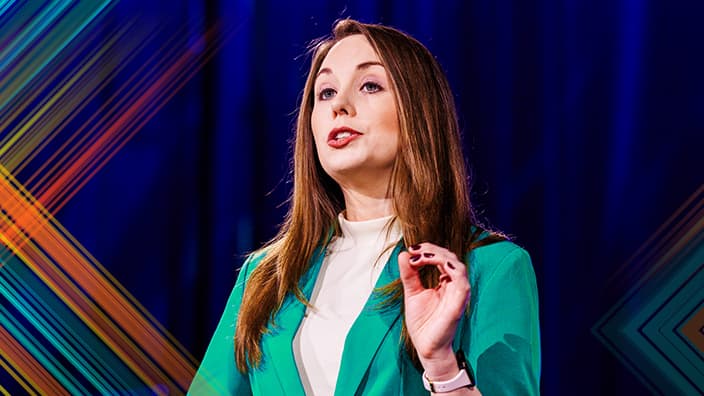 Head and shoulders portrait of a presenter who has long brunette hair wearing a teal green blazer with their hand raised in emphasis