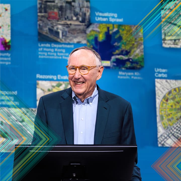 Esri president Jack Dangermond speaking onstage with user-submitted images and maps in the background