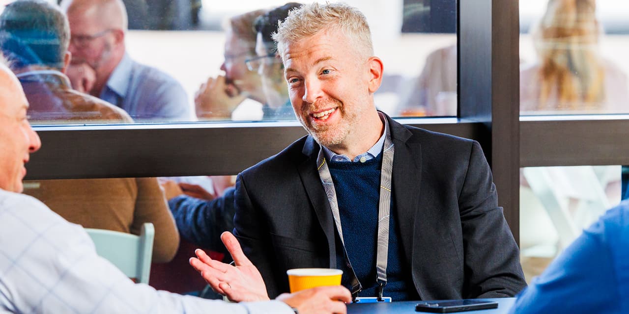 Conference attendees seated in groups talking; one of them smiles and gestures with one hand 