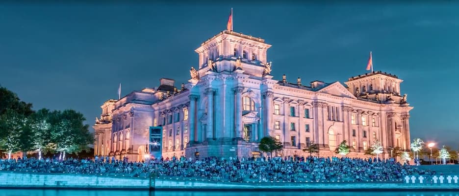 The iconic Reichstag building in Berlin illuminated at night as a large crowd sits on a gentle slope in front of the building