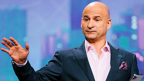 Presenter in a dark suit and light purple dress shirt with their hand raised in emphasis against a pastel blue and purple backdrop