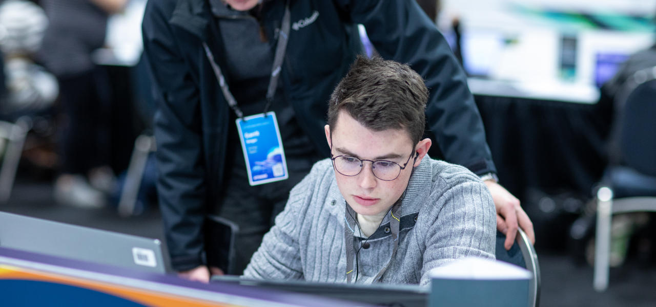 An event attendee seated at a table using a computer as another person leans over them to help