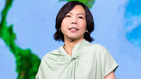 Presenter wearing a mint green blouse against a brightly lit blurred map backdrop